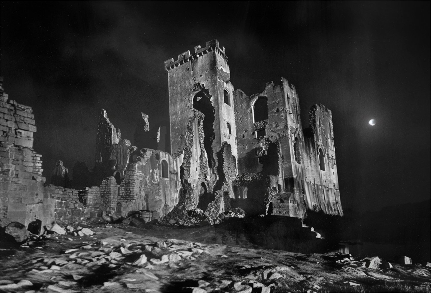 Black and white photo with film grain depicting an ancient medieval style castle on a deserted landscape at night with a bright moon.